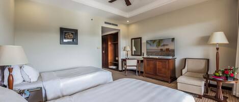 Habitación de hotel con dos camas, un escritorio, una silla, un ventilador de techo, una lámpara y un cuadro en la pared.