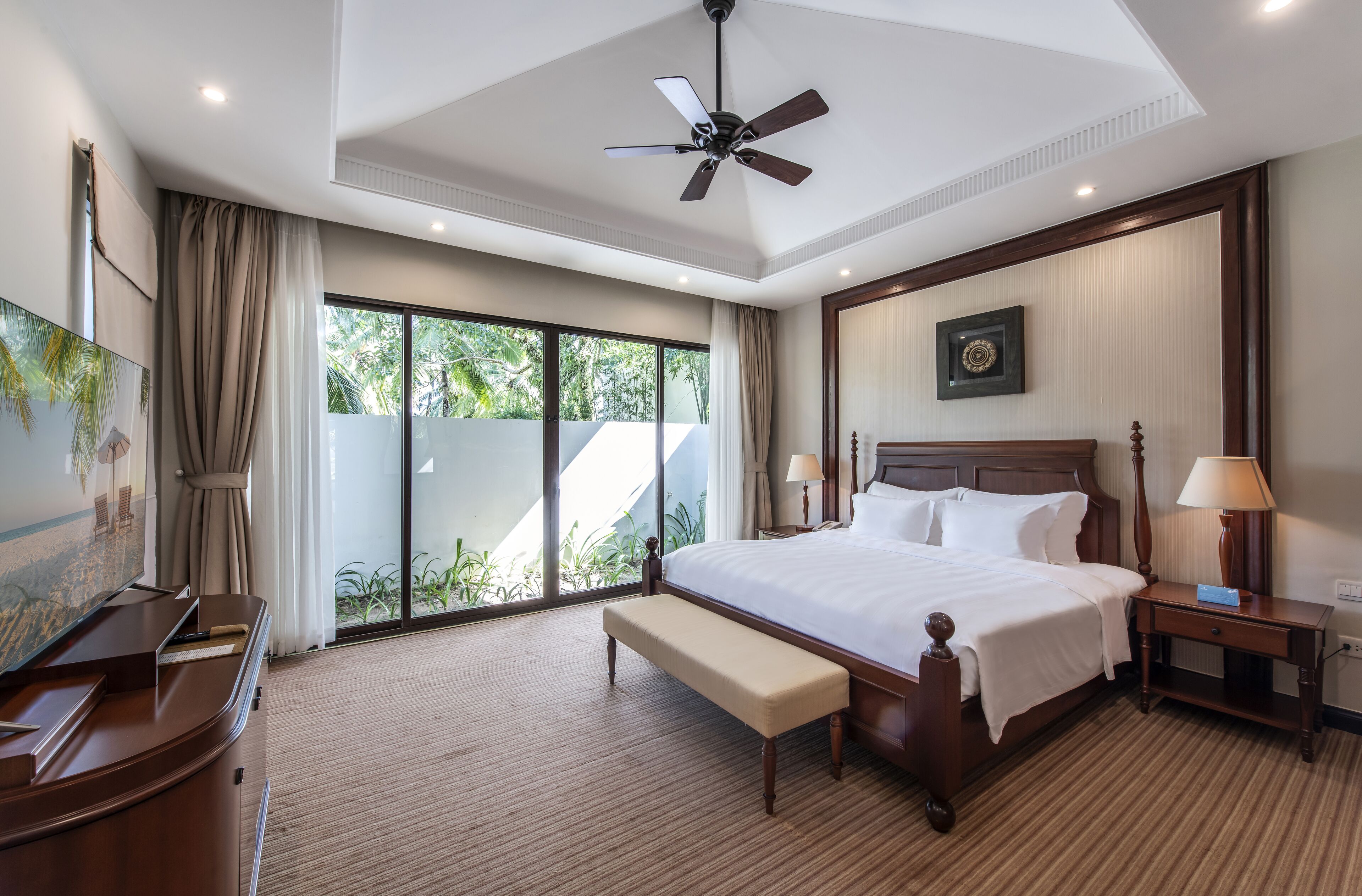 A hotel room with a large bed, a desk, a bench, a ceiling fan, and a view of greenery through the window.