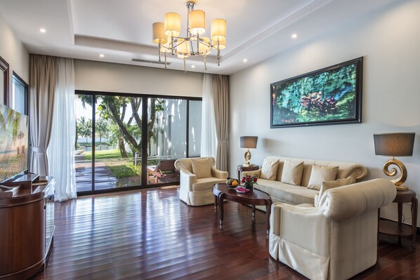 A spacious living room with a large sofa, two armchairs, a coffee table, and a television. There is a large window with curtains, a chandelier, and a painting on the wall.