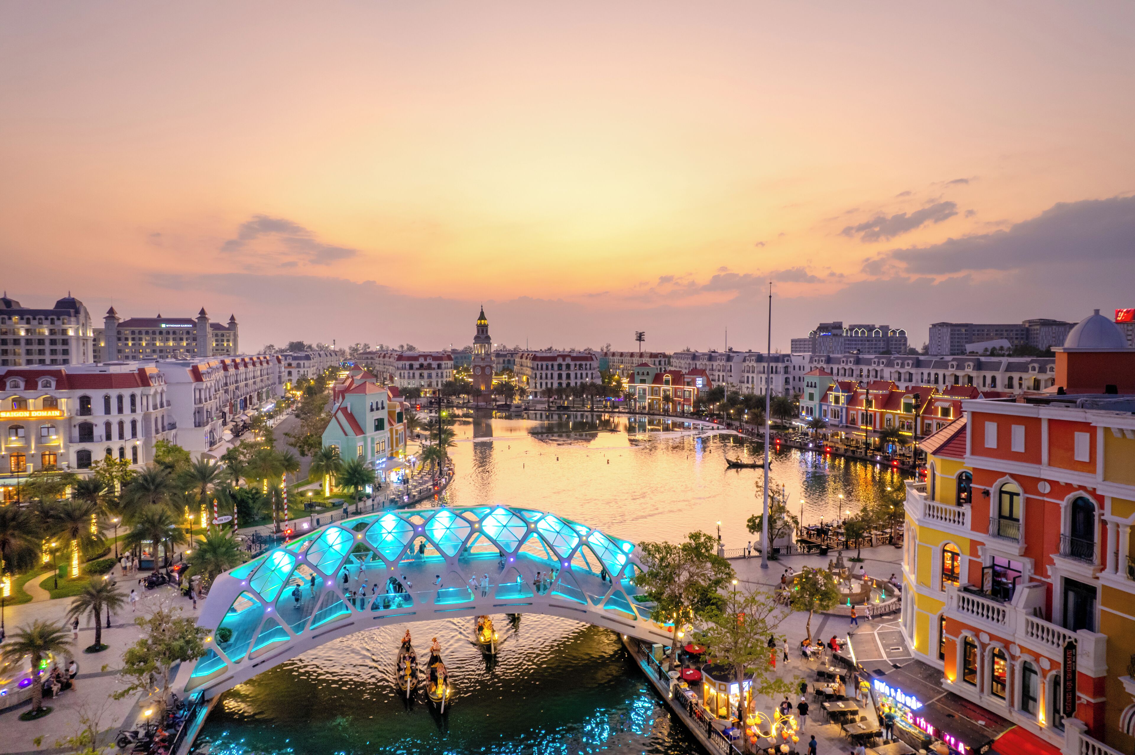 A riverside promenade with a glass bridge, historic buildings, and a view of the city skyline at sunset.
