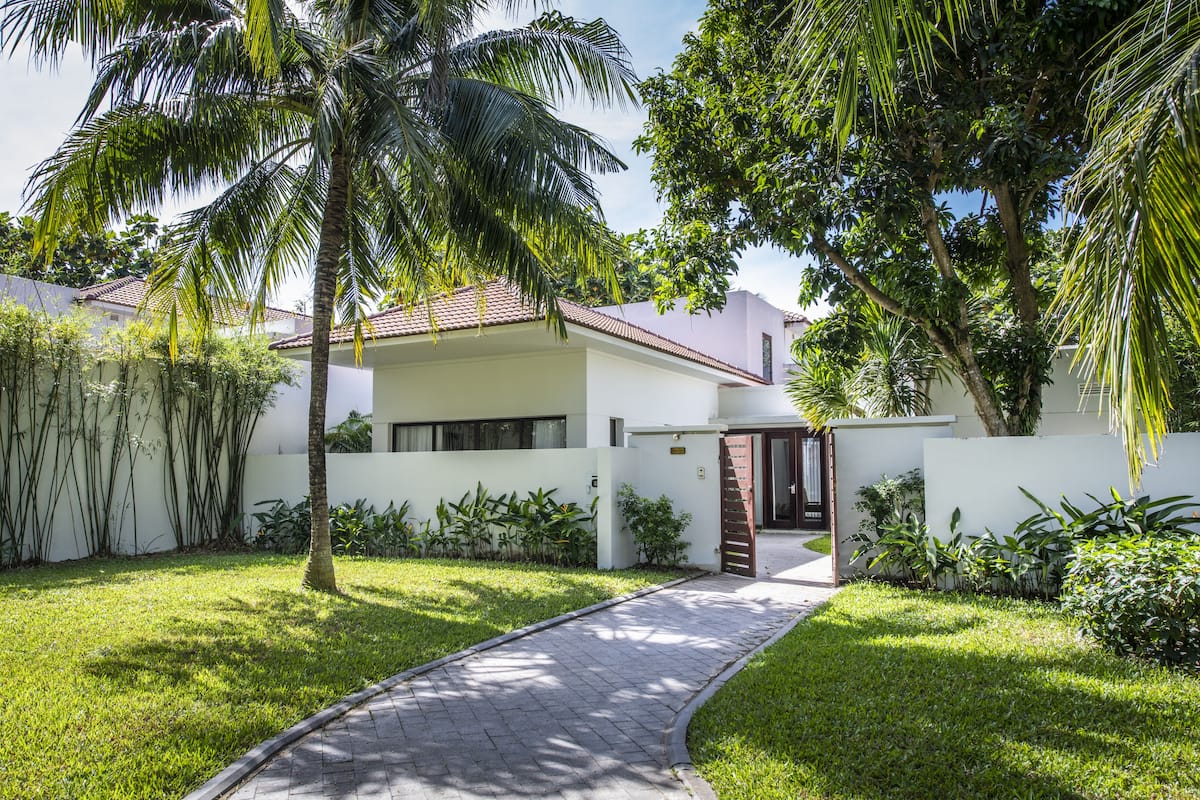 A modern house with a paved driveway, surrounded by lush greenery and palm trees.