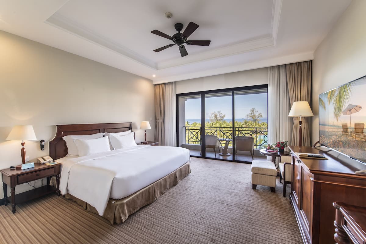 A hotel room with a large bed, a desk with a lamp, a chair, a balcony with a view, and a ceiling fan.