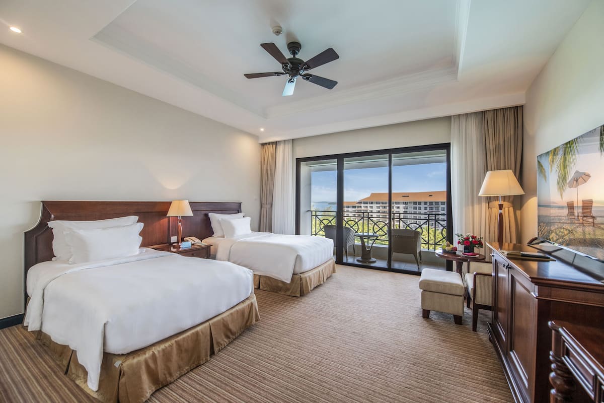 A hotel room with two beds, a desk, a chair, a balcony with a view, and a ceiling fan.