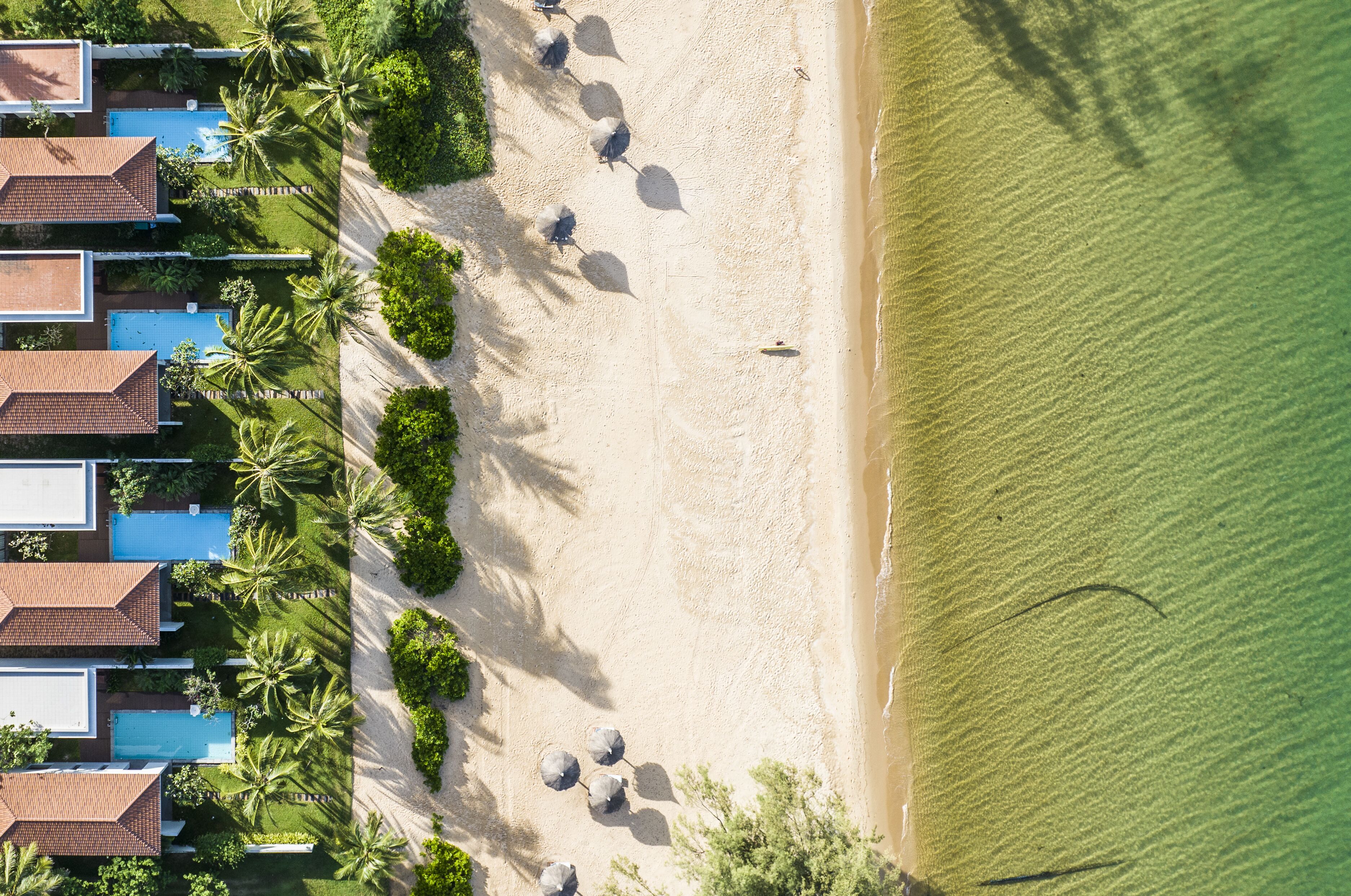 An aerial view of a beachside resort with multiple bungalow-style buildings, a sandy beach, and palm trees.