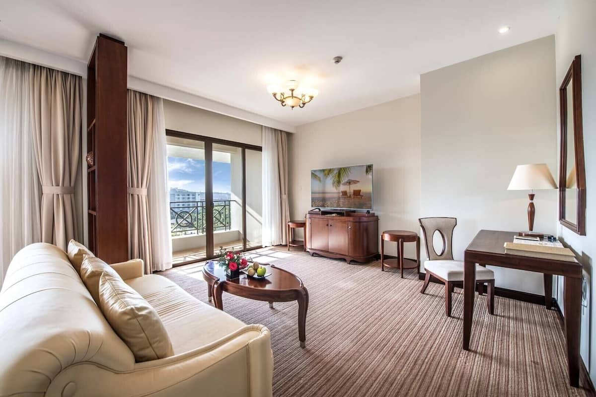 A hotel room with a sofa, a coffee table with fruit, a TV, a desk with a lamp, and a balcony with a view.