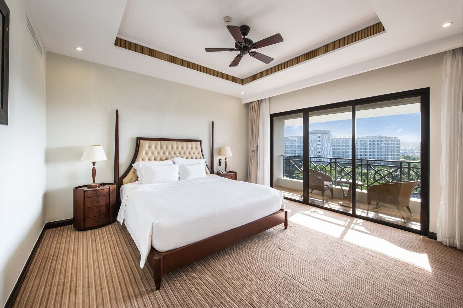 A hotel room with a large bed, a ceiling fan, a nightstand with a lamp, and a balcony with outdoor furniture.
