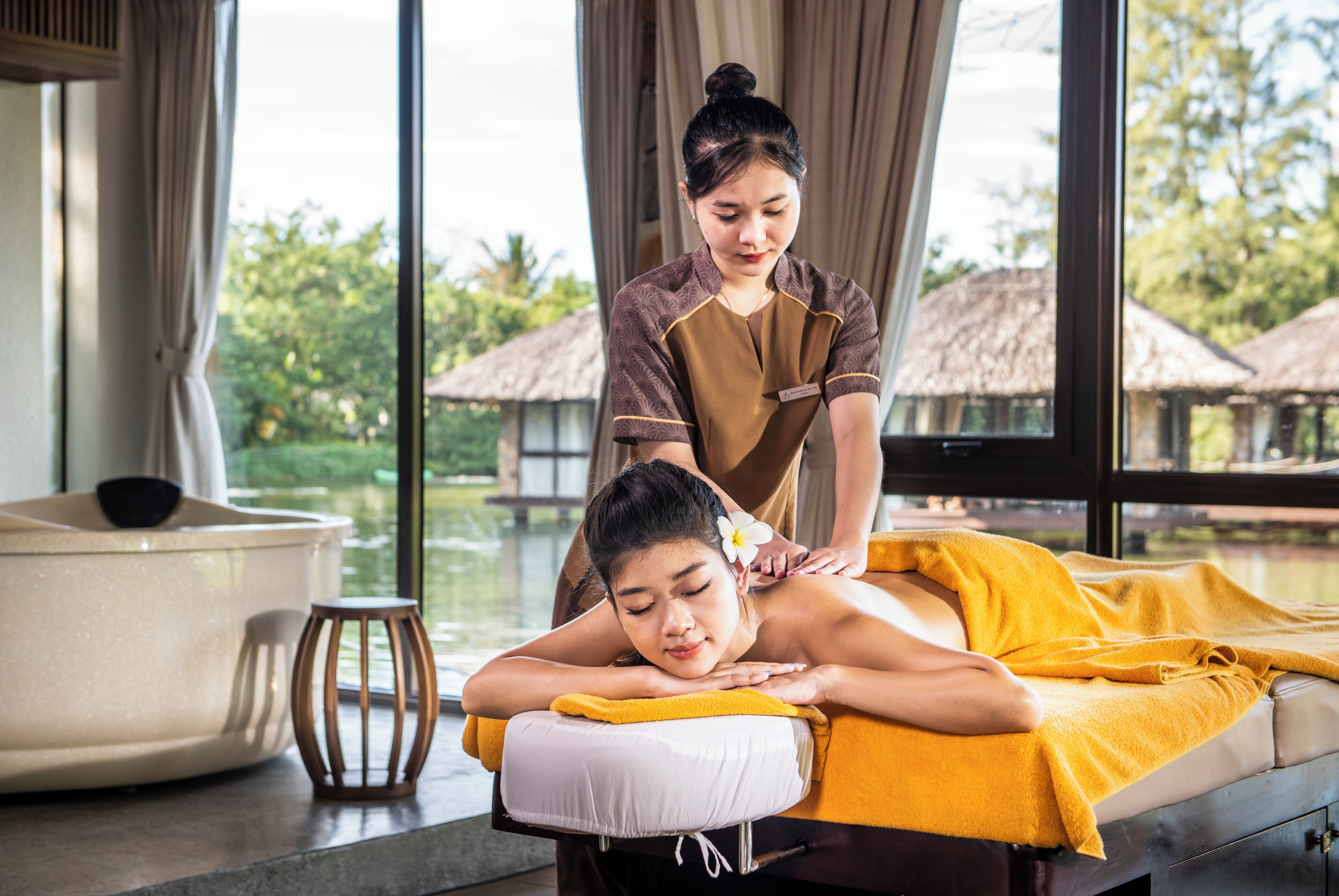 A massage therapy room with a massage table, a massage therapist, and a large window with a view of a thatched-roof structure and greenery.