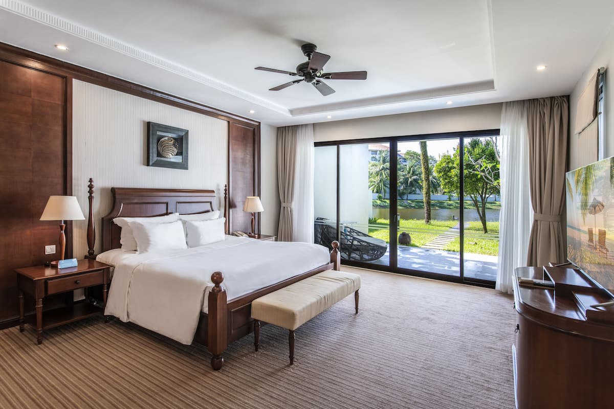A spacious hotel room with a large bed, a ceiling fan, a view of the outdoors, and a TV on a wooden cabinet.