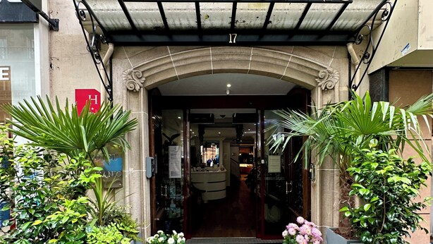 L'entrée de l'hôtel Oceanic, avec son auvent en verre, un grand « H » rouge et des plantes en pot.