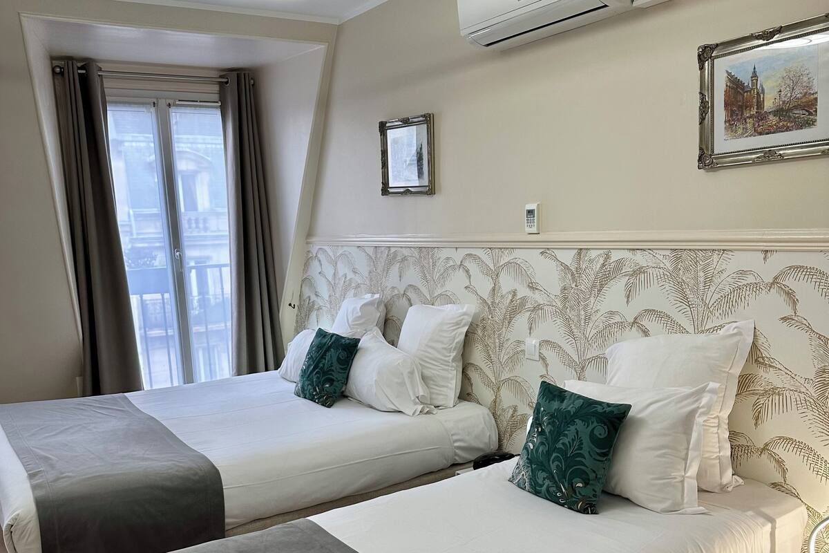 A hotel room with two beds, a modern chandelier, an air conditioning unit, a framed picture, and a window with curtains.