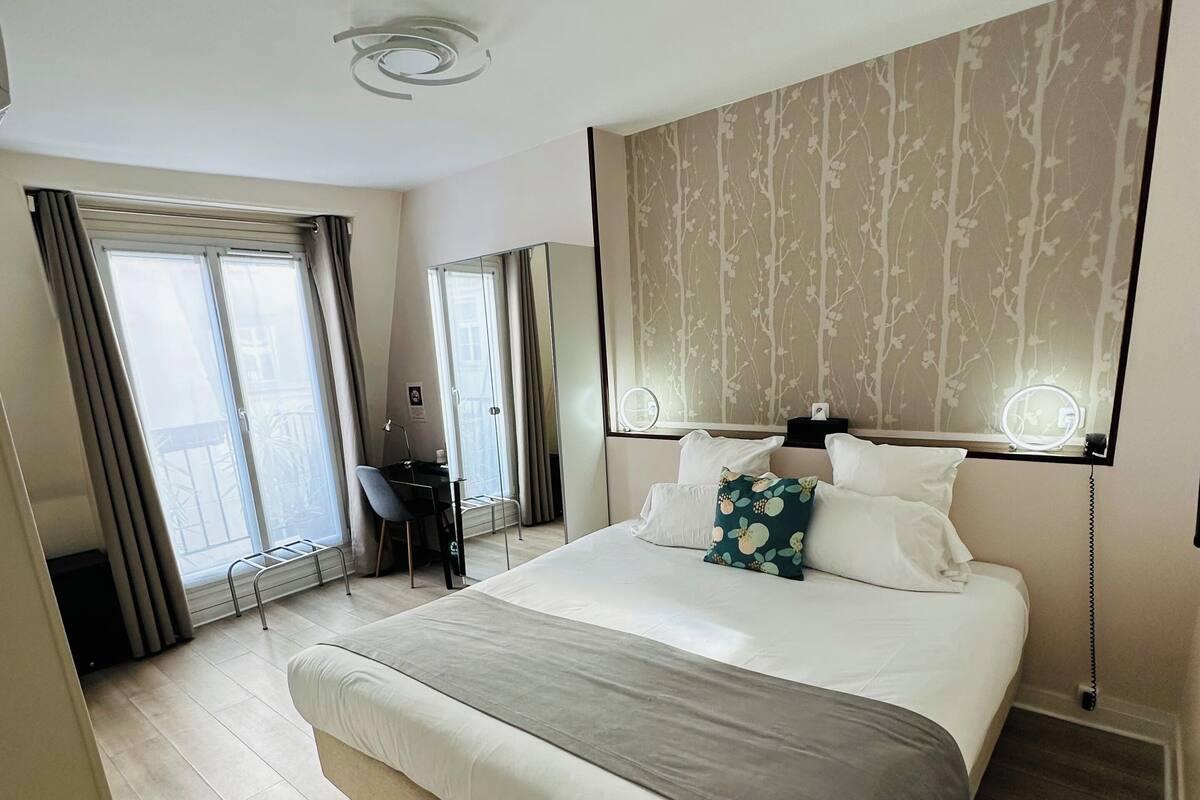 A hotel room with a large bed, a desk, a chair, a window with curtains, and a wall-mounted light fixture.