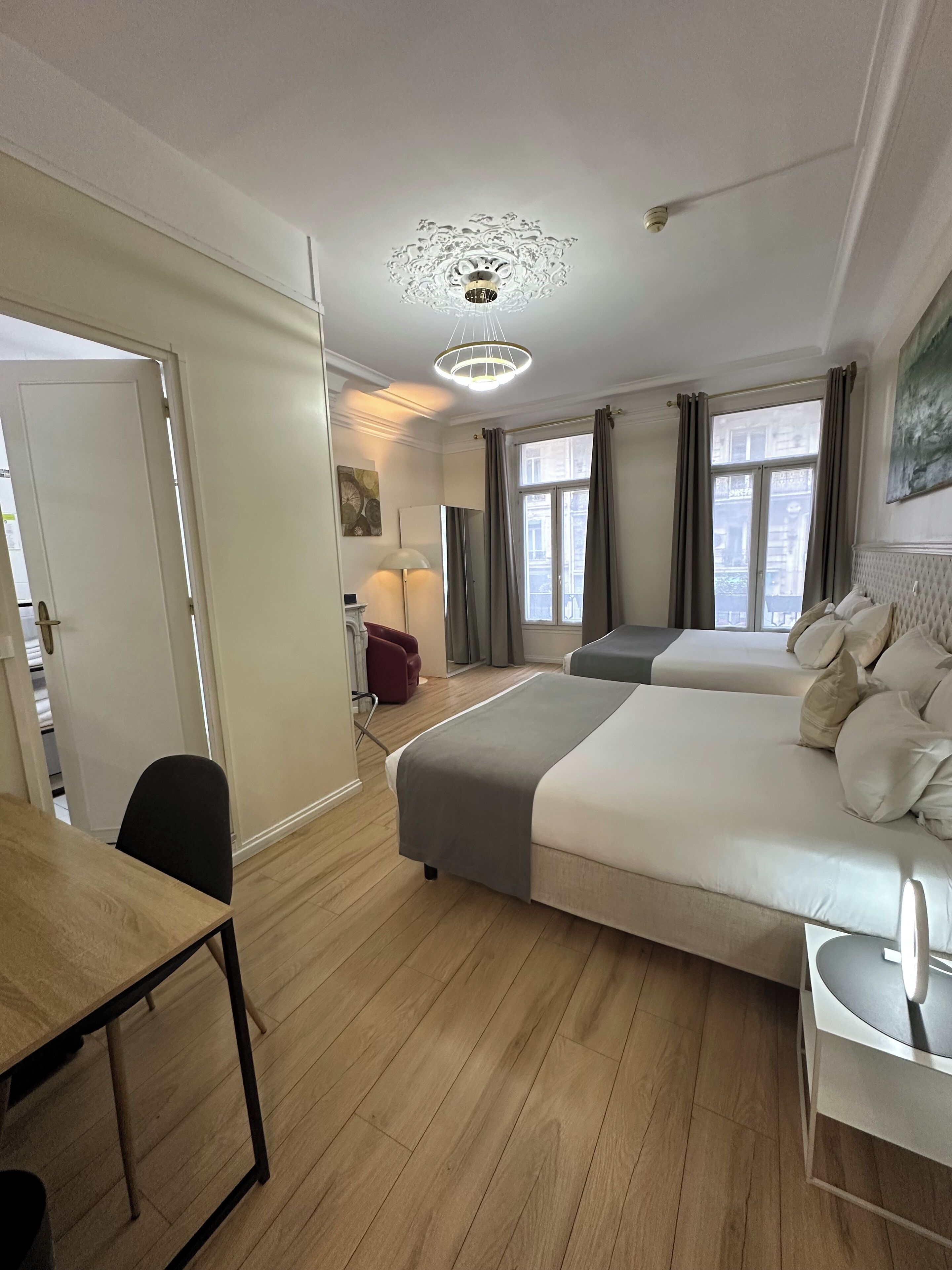 A hotel room with two beds, a desk, a chair, a lamp, and a view of the city through the window.