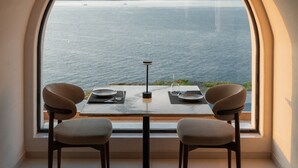 Un coin repas comprenant une table et deux chaises, une grande fenêtre en arc de cercle offrant une vue sur la mer et les montagnes, et un plafond au revêtement en bois.