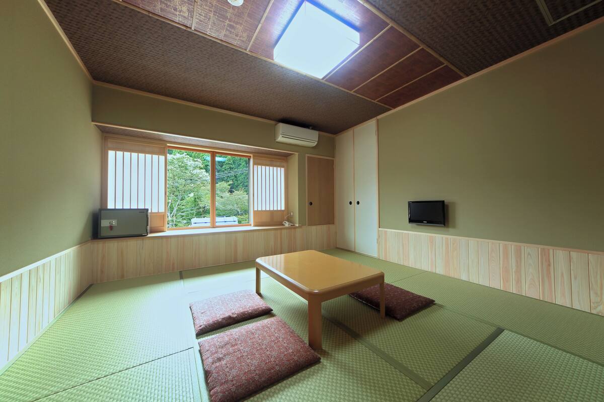 Una habitación con suelo de tatami, una mesa de madera baja y cojines. Hay una ventana con persianas, un televisor en la pared y un aire acondicionado.