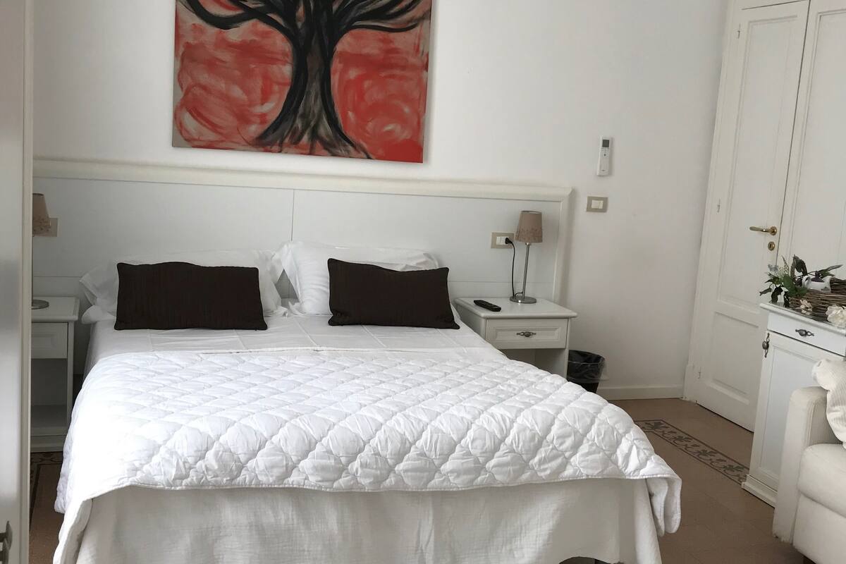 Une chambre à coucher bien rangée, avec un grand lit, une table de chevet avec une lampe, une commode et un tableau accroché au mur.