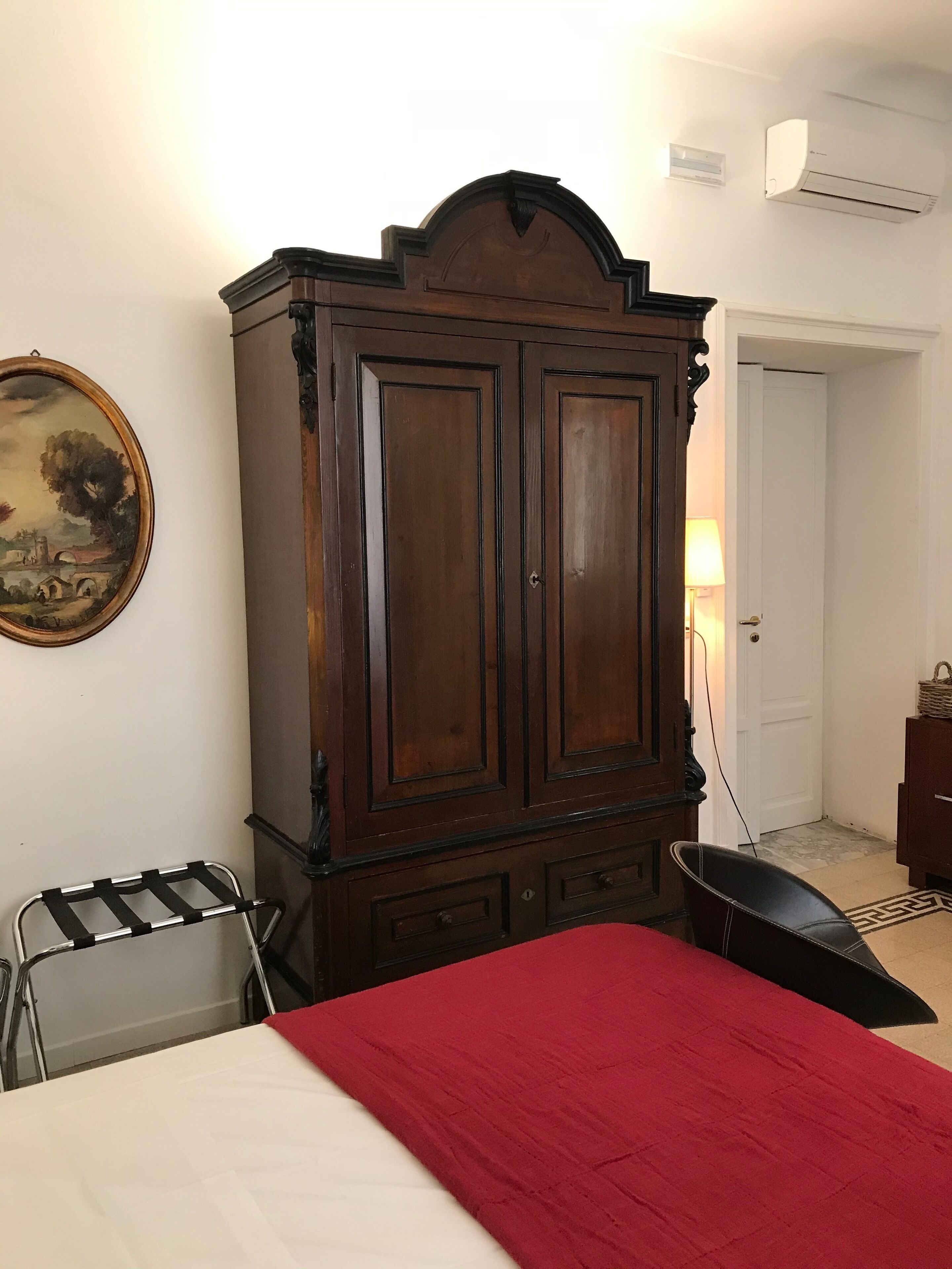 Une chambre avec une grande armoire en bois, un lit recouvert d’une couverture rouge, une chaise, une lampe et un tableau accroché au mur.