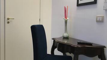 Une pièce avec un fauteuil bleu foncé, une petite table sur laquelle se trouvent un vase et une télécommande, un tableau encadré accroché au mur et une porte.