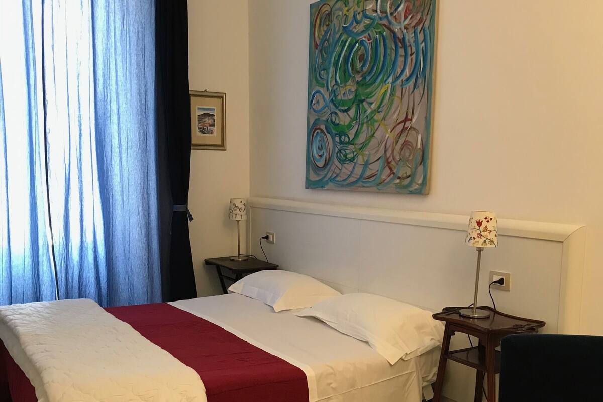 Un lit bien fait, recouvert d’une couverture rouge et blanche, une table de chevet avec une lampe, et un tableau abstrait aux couleurs vives accroché au mur.