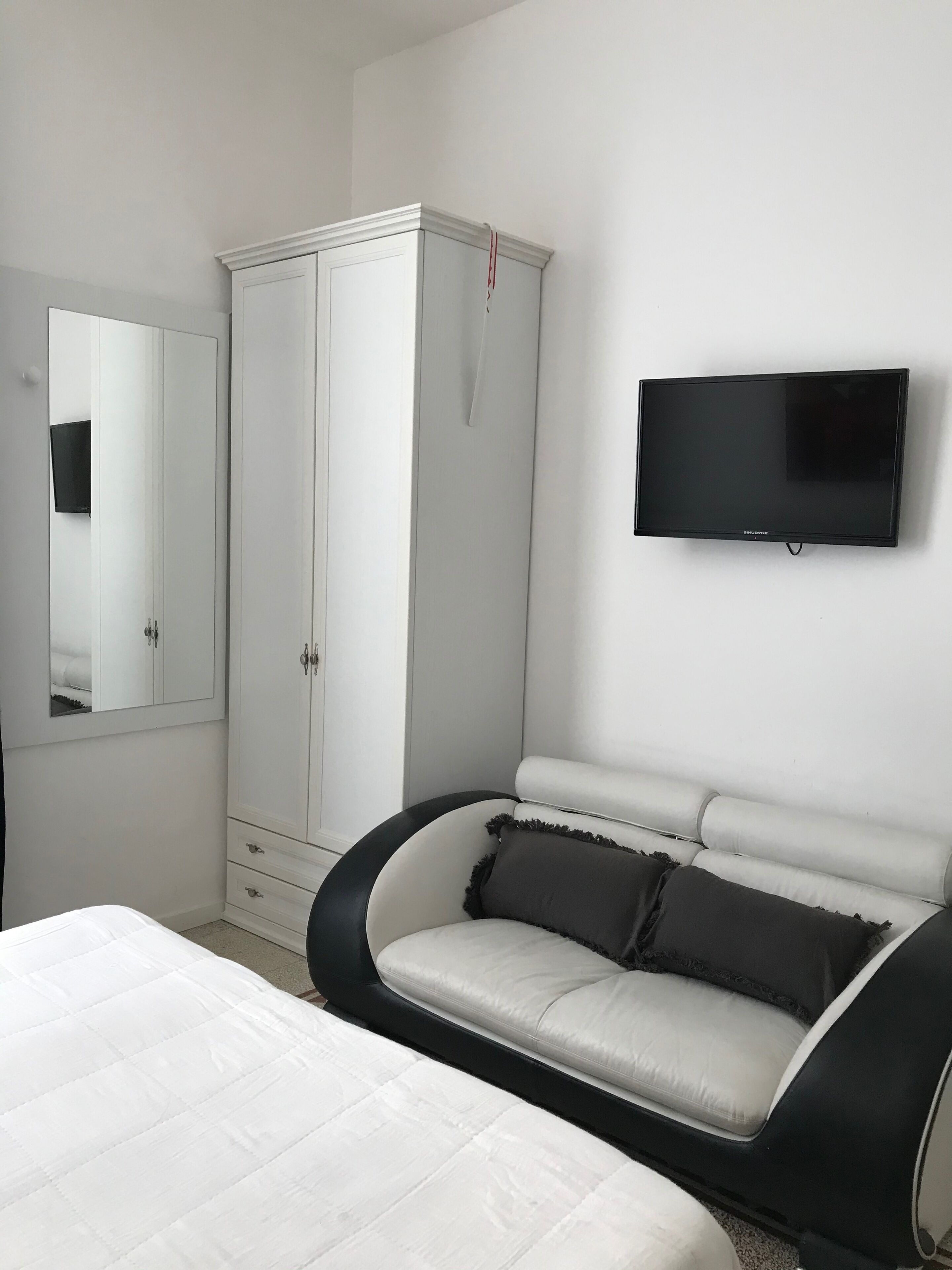 A bedroom with a bed, a white wardrobe, a television on the wall, and a black and white sofa.