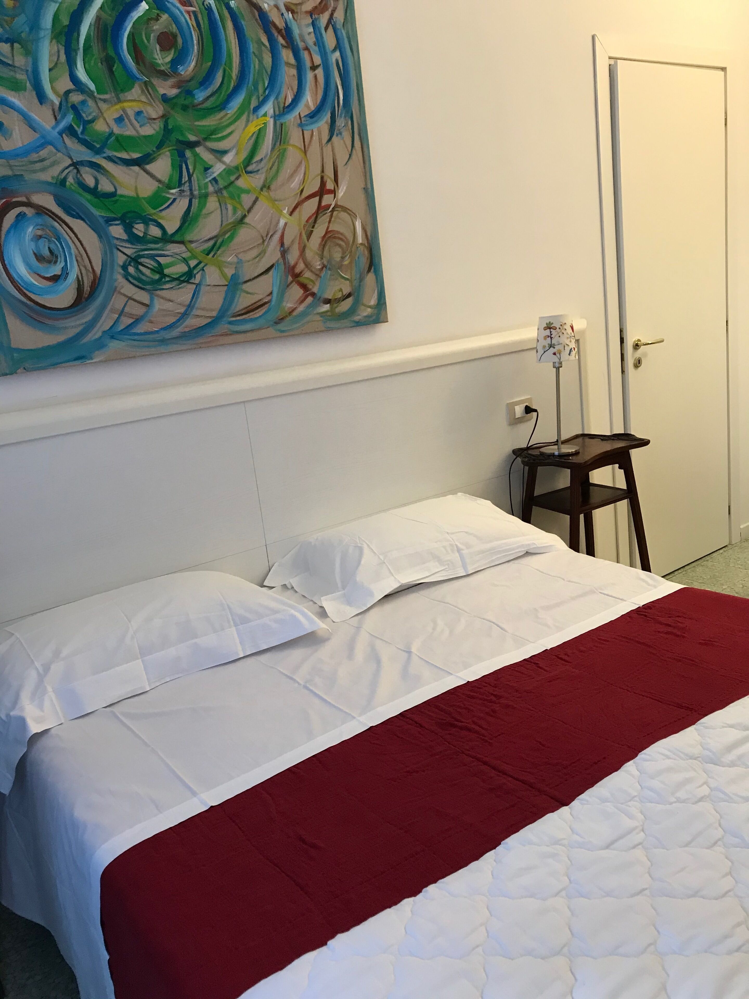 Una cama bien hecha con ropa de cama blanca y roja, un taburete de madera pequeño y un cuadro abstracto y colorido en la pared.