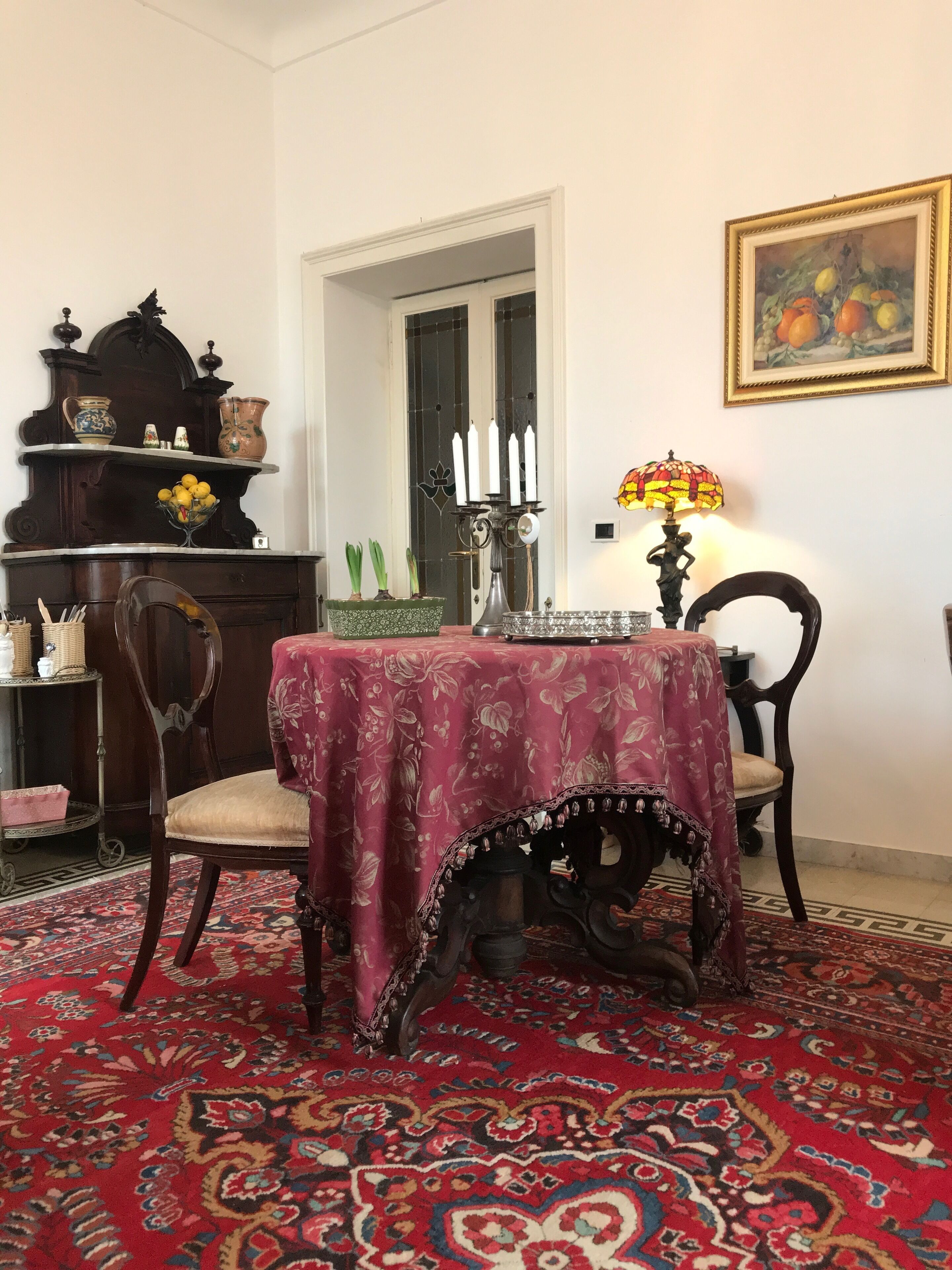A dining area with a table covered in a floral tablecloth, surrounded by ornate chairs. A chandelier hangs above the table, and a lamp with a stained glass shade is on the table.