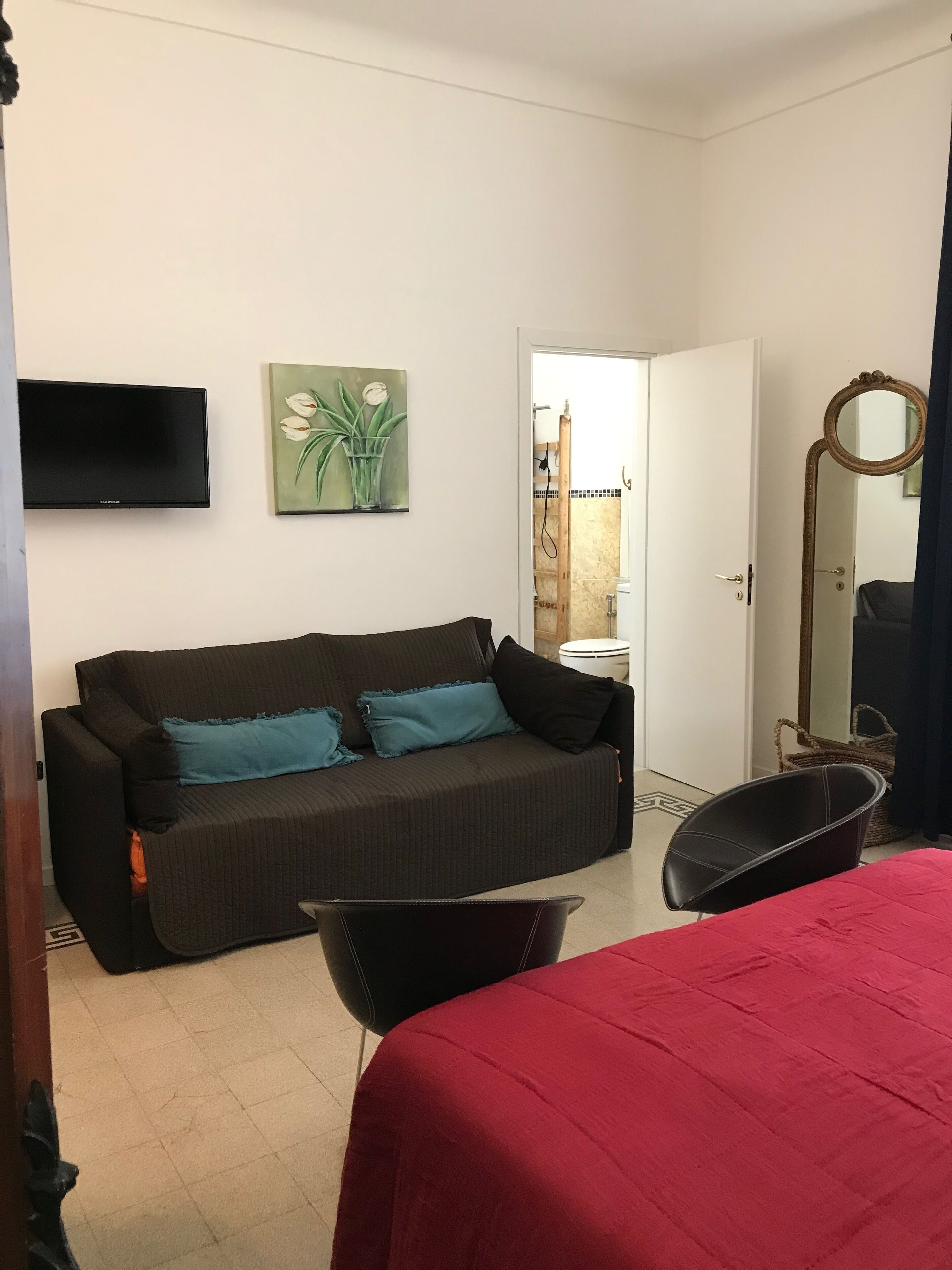 Ein Zimmer mit einem dunkelgrauen Sofa, einem roten Bett, einem schwarzen Sessel, einem Fernseher, einem Pflanzenbild und einem durchgehenden Spalt zu einem Badezimmer.