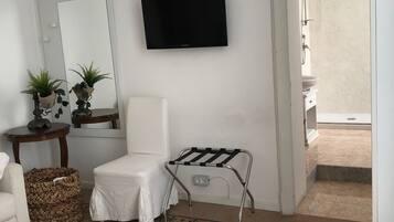 A bedroom with a bed, a chair, a small table with plants, a TV mounted on the wall, and a view of the outside balcony.