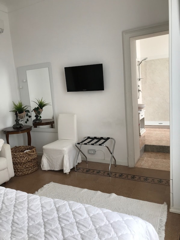 A bedroom with a bed, a chair, a small table with plants, a TV mounted on the wall, and a view of the outside balcony.