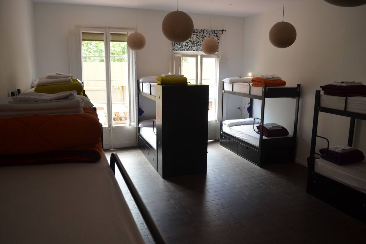 A dormitory room with bunk beds, a desk, and a window overlooking a balcony.