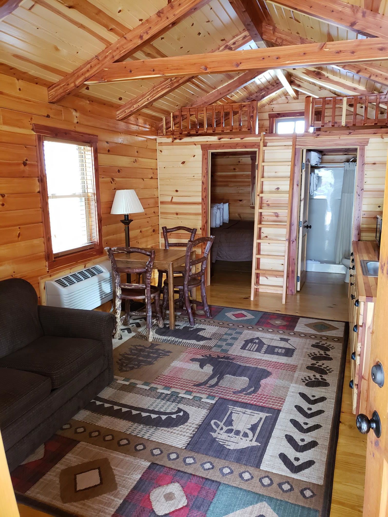 Interior acogedor de una cabaña con comedor, un sofá, un tapete estampado y una escalera que lleva a un dormitorio.