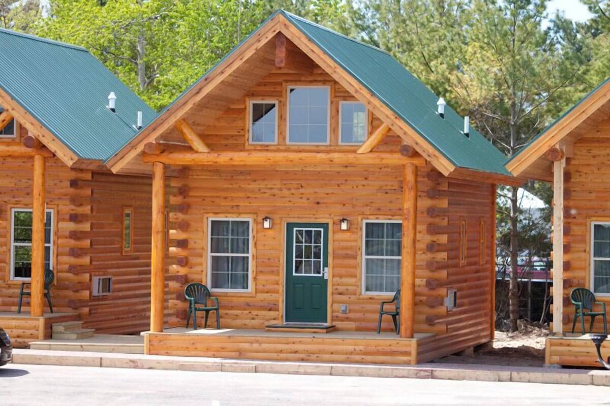 Une cabane en bois avec un toit vert, deux fenêtres à l’avant, une porte verte et une véranda avec deux chaises.