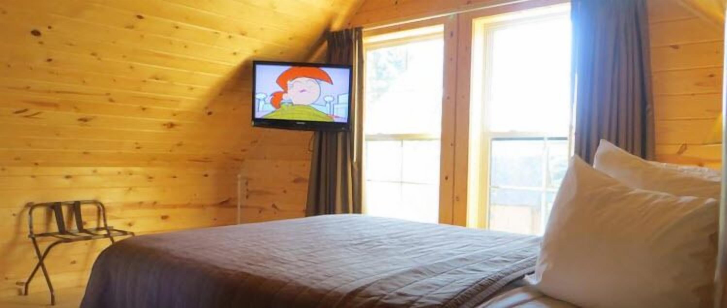 A wooden cabin room with a bed, a television on the wall, and a window.