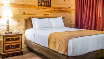 A wooden-paneled room with a bed, a nightstand with drawers, a lamp, and a framed picture on the wall.