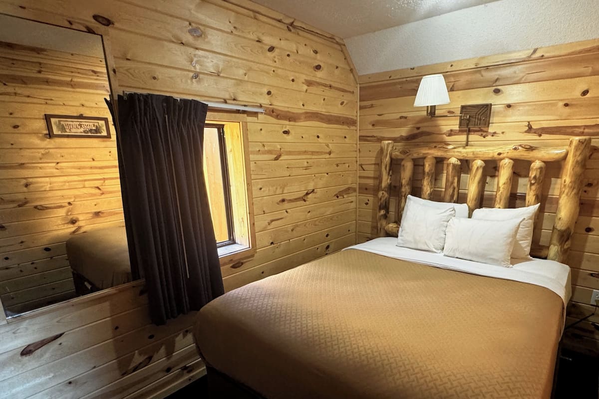 Une pièce de style cabane en bois, avec un lit, une fenêtre avec des rideaux, une lampe fixée au mur et un tableau encadré.