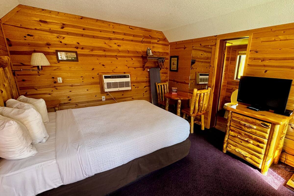 Une pièce de cabane en bois comprenant un lit, un téléviseur posé sur une commode en bois et une petite table avec des chaises.