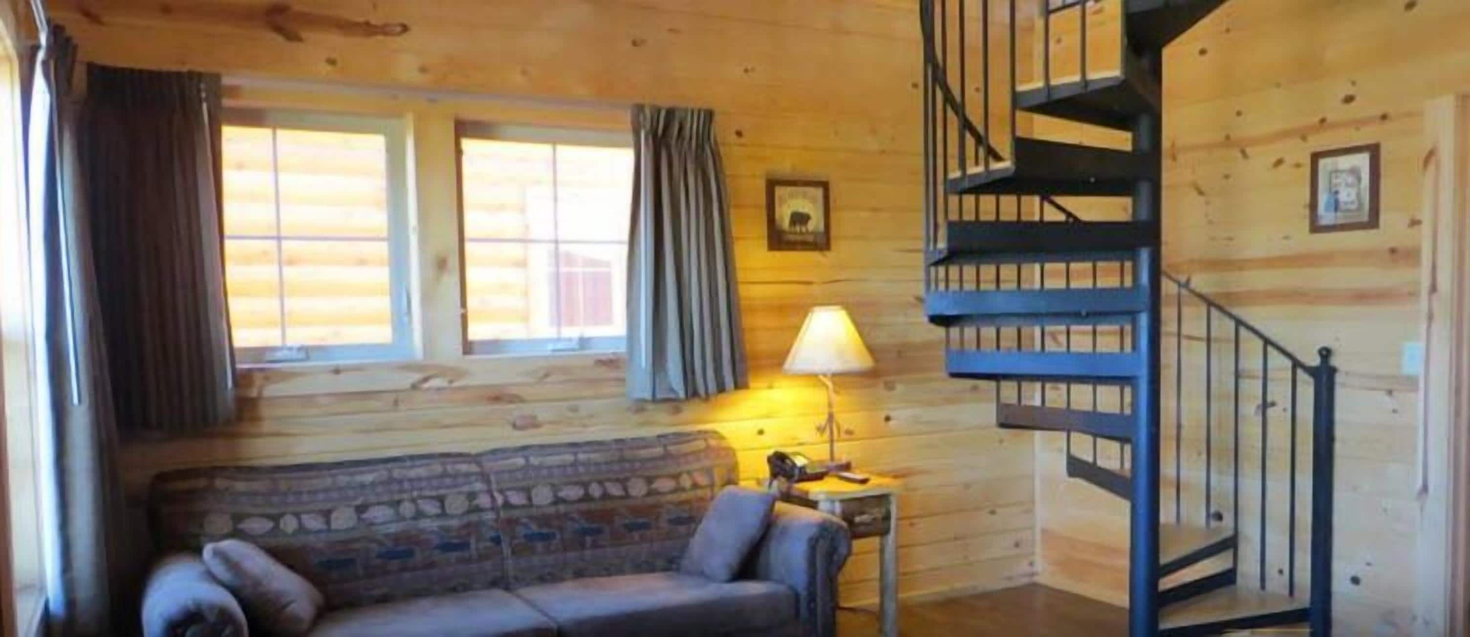 A log cabin interior with a stone-patterned sofa, a lamp, a staircase, and wooden walls.