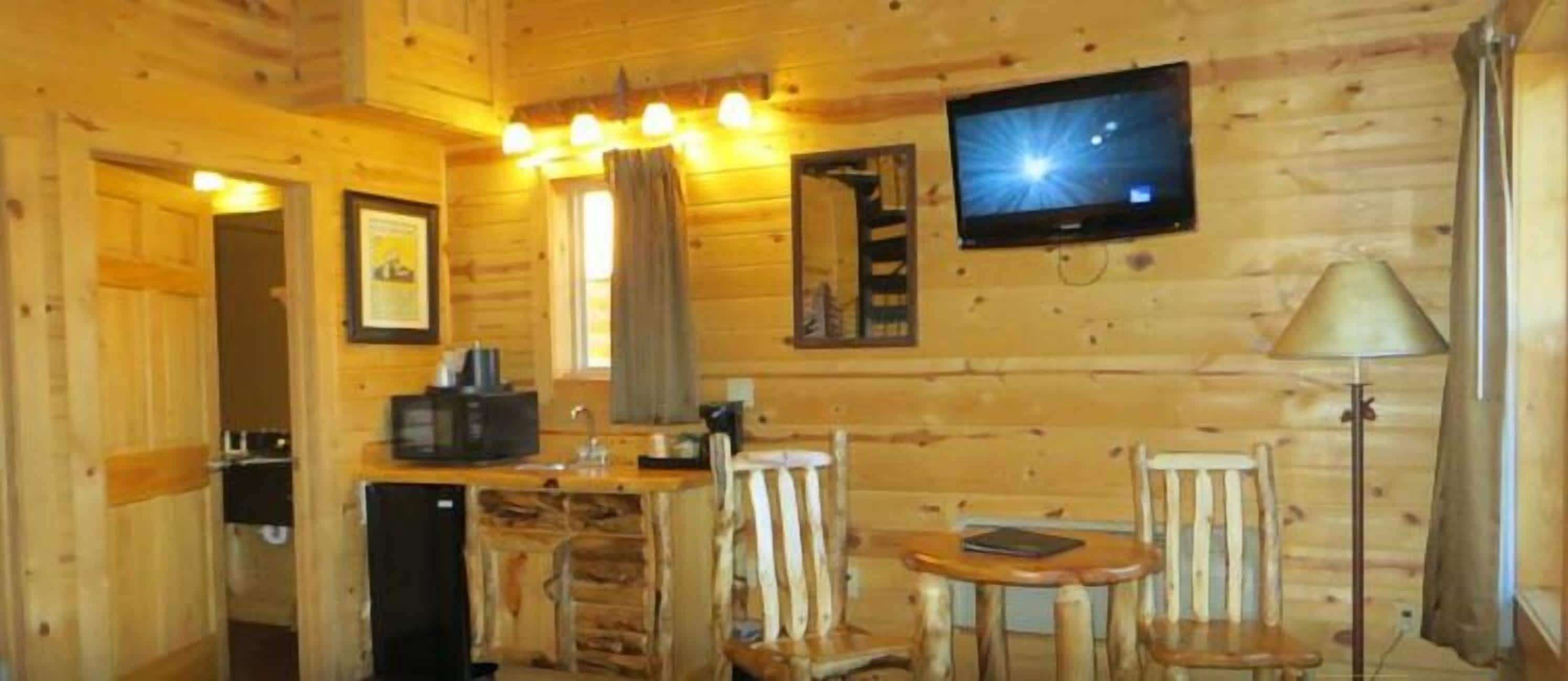 A wooden cabin interior with a kitchen area, a dining table with chairs, a television mounted on the wall, and a lamp.