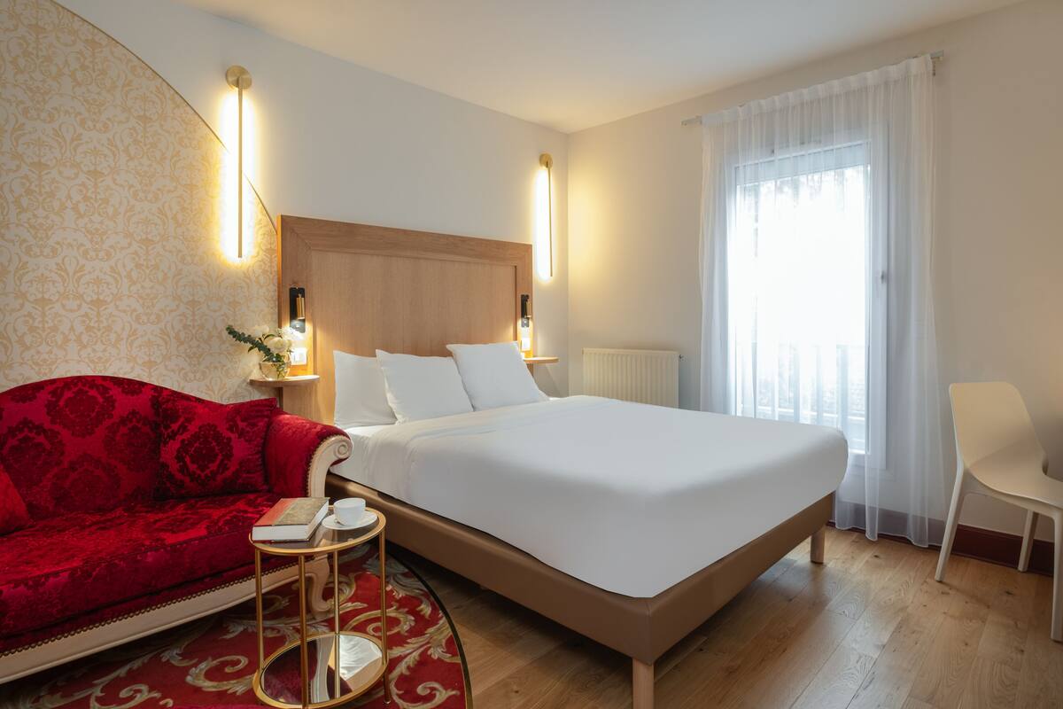 A hotel room with a large bed, a red sofa, a small round table, and a chair.