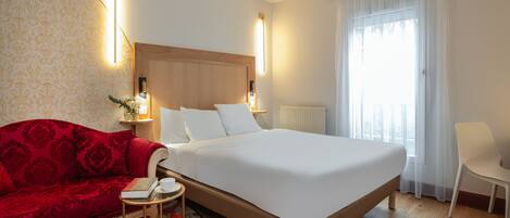 A hotel room with a large bed, a red sofa, a small round table, and a chair.