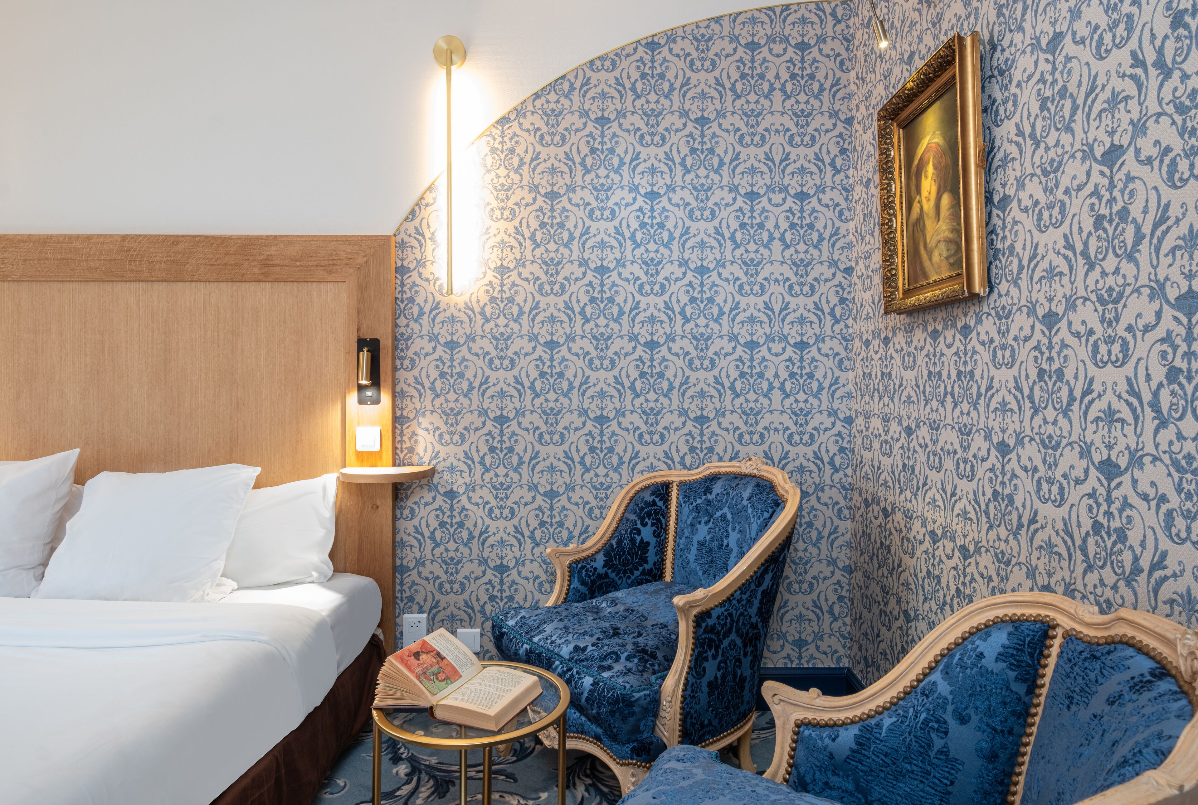 A hotel room with a bed, two blue upholstered chairs, a small round table with a book, and a wall with a patterned wallpaper and a framed picture.