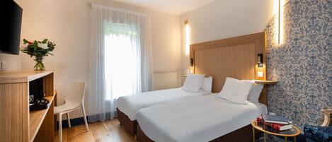 A hotel room with two beds, a desk, a chair, a TV, and a bedside table with books.