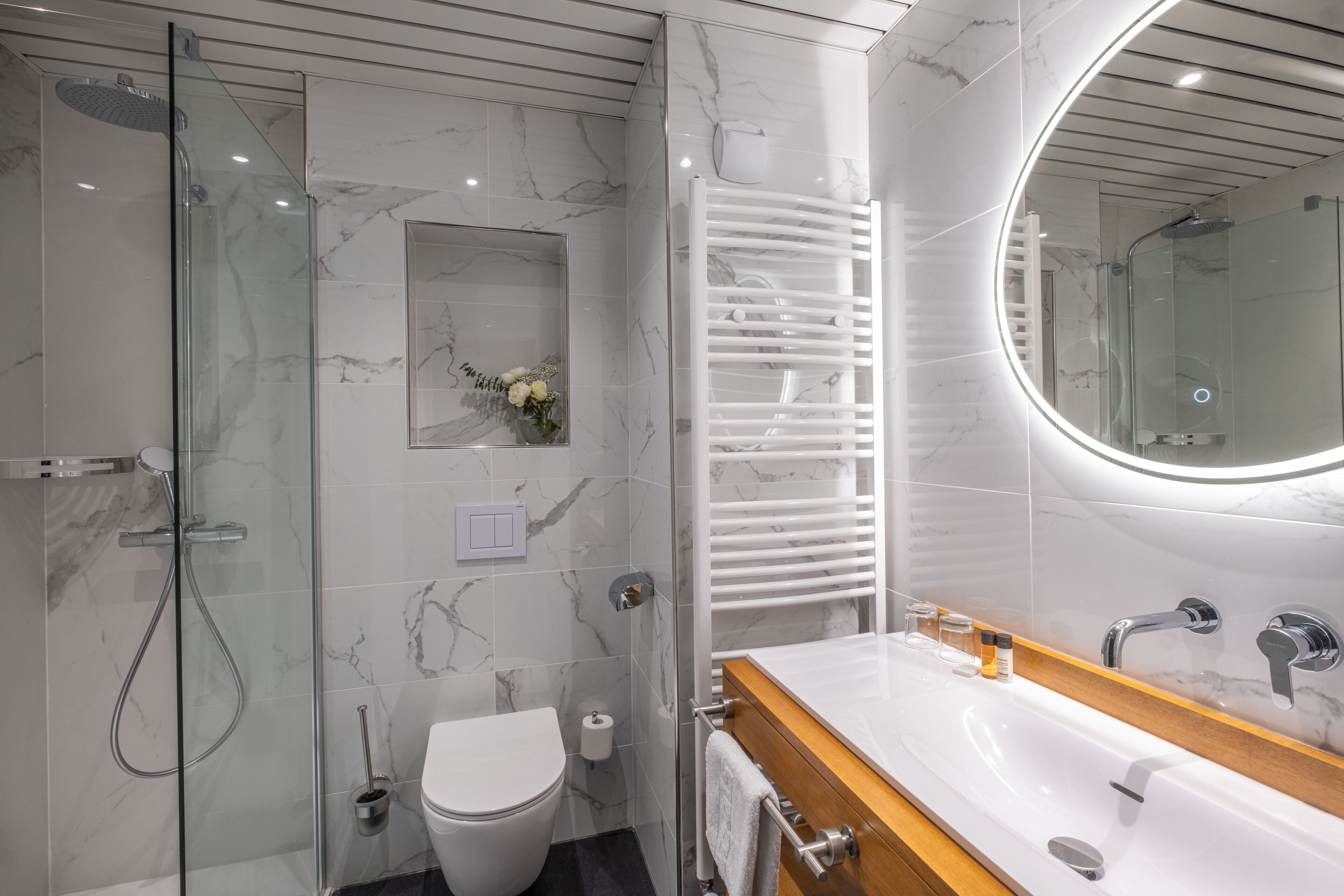 Un baño moderno con ducha de vidrio, inodoro blanco, tocador con espejo y perchero para toallas.
