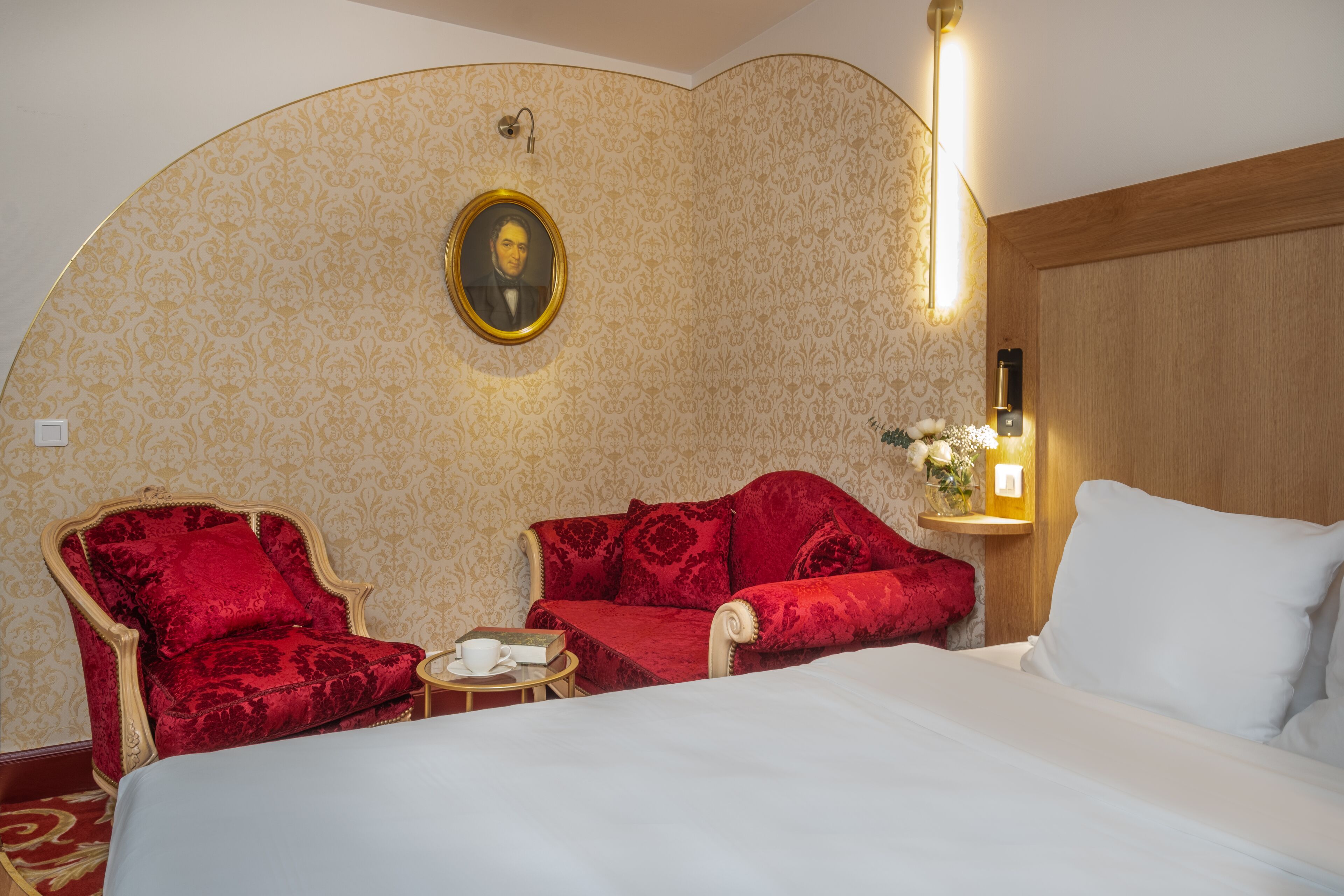 Une chambre d’hôtel avec un grand lit, deux fauteuils rouges, une petite table ronde avec une tasse et un livre, un tableau accroché au mur et une table de chevet avec une lampe et des fleurs.