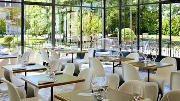 A restaurant with tables and chairs arranged in a spacious, sunlit interior with large windows offering a view of a garden and a building.