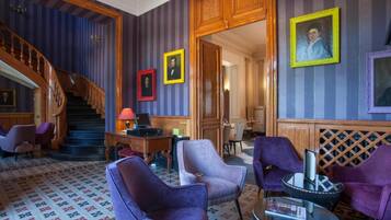 A lobby with a staircase, purple armchairs, a wooden desk, and framed portraits on the wall.