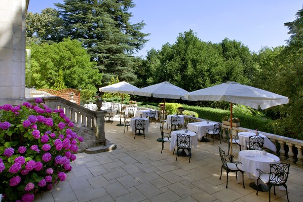 An outdoor patio with tables and chairs, surrounded by greenery and trees.