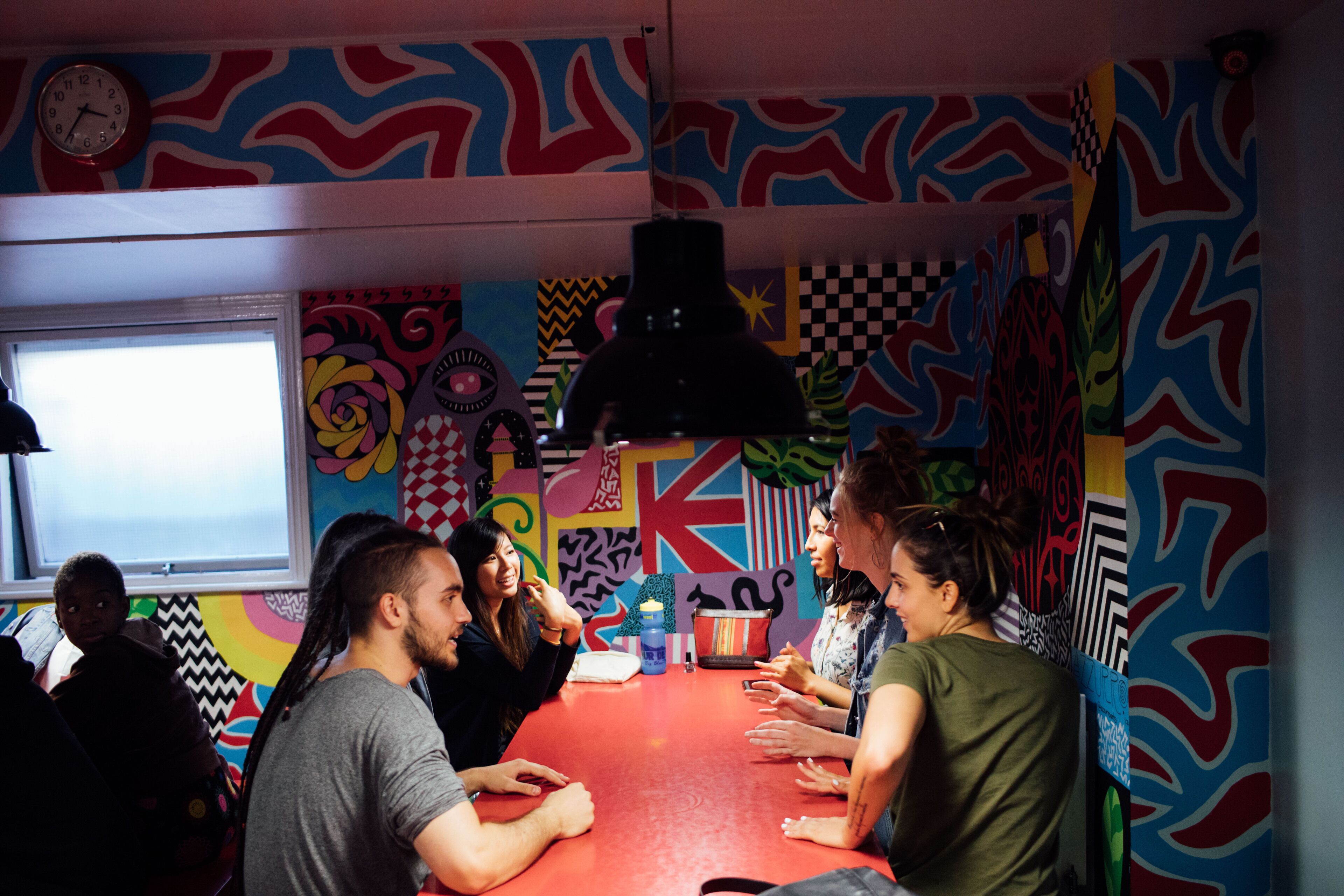 Een groep mensen zit rond een rode tafel in een kamer met kleurrijke, abstracte muurkunst.