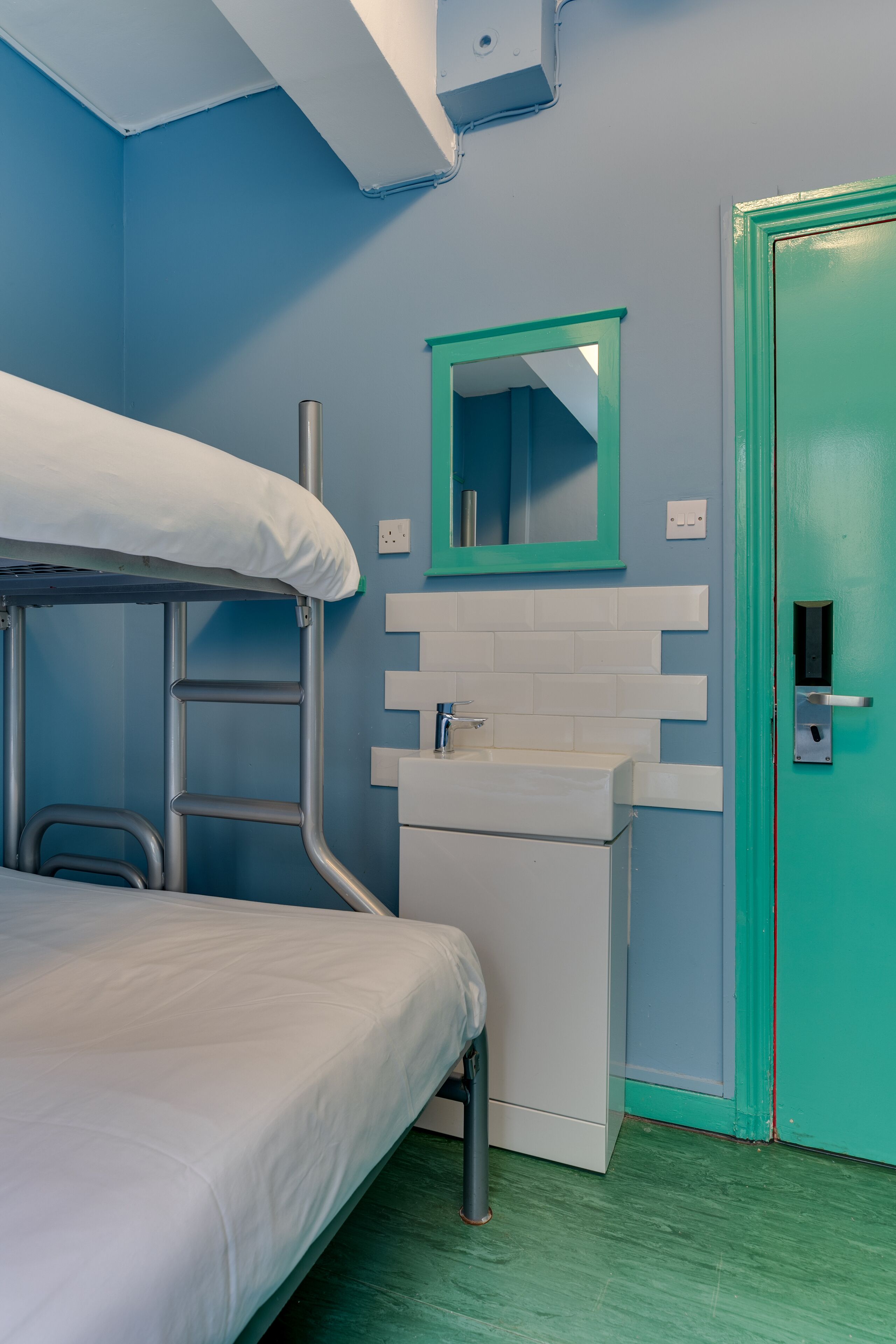 Una cama litera con colchón blanco, un lavamanos blanco con espejo arriba y una puerta verde azulado.