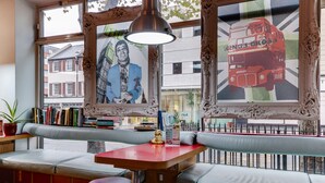 A cozy seating area with a red table, cushioned benches, and a large window with artwork and a view of buildings. - Clink 261 Hostel (London)