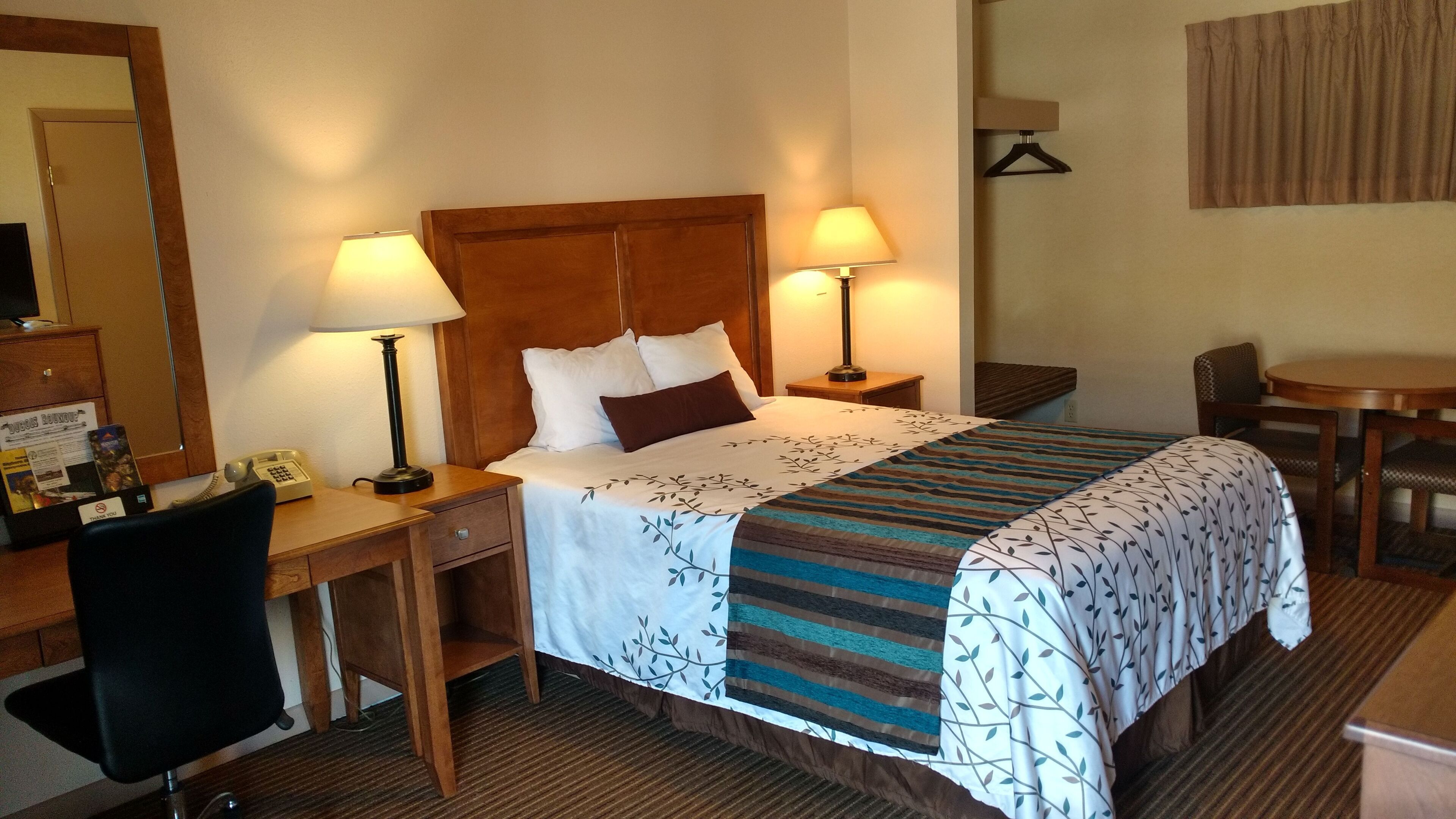 A hotel room with a bed, two bedside lamps, a desk with a chair, a small table, and a hanging coat rack.